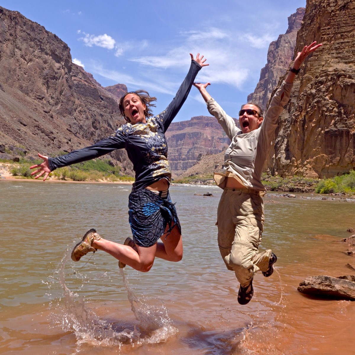 Two people jump for joy out of shallow water near the shore of the Colorado River during a sunny summer day