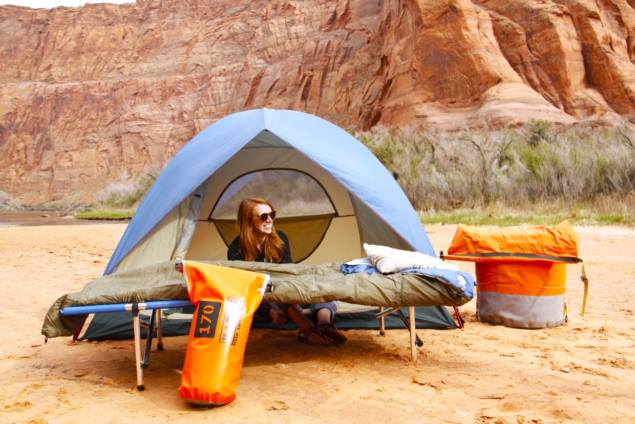 A women sit inside of a tent with the door open and with river rafting camping gear around her, including a cot, sleeping bag, pillow, and two large dry bags by Hatch River Expeditions