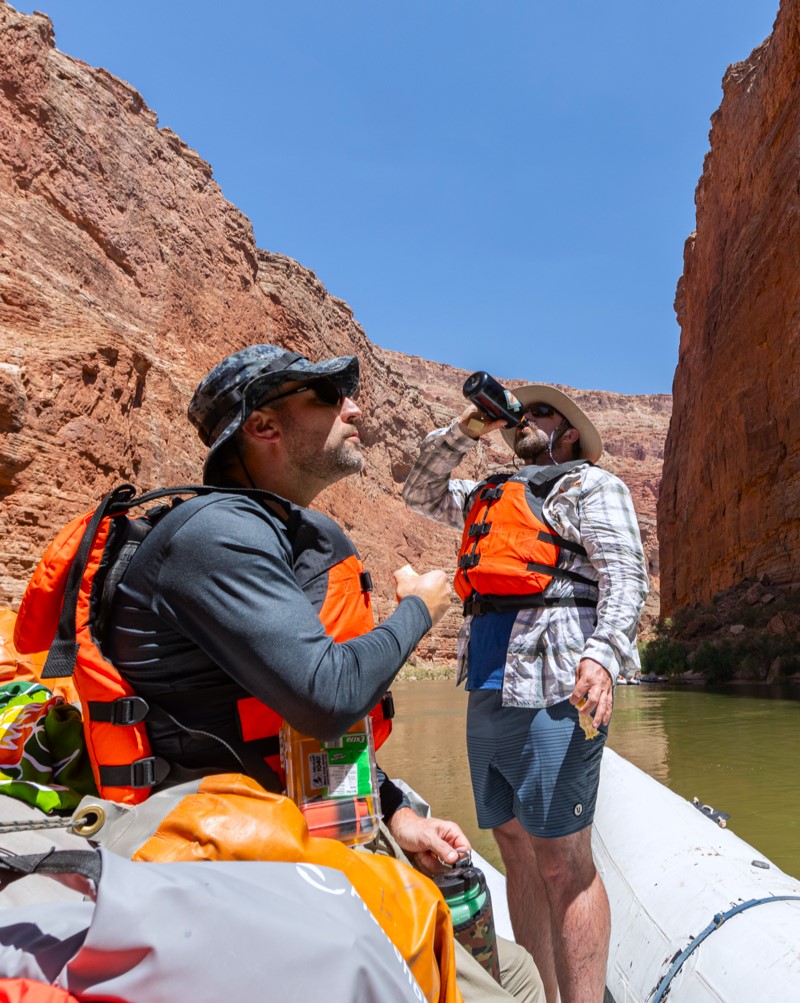 two men wearing life vests on a river raft in Grand Canyon who are looking at the cliffs while drinking water