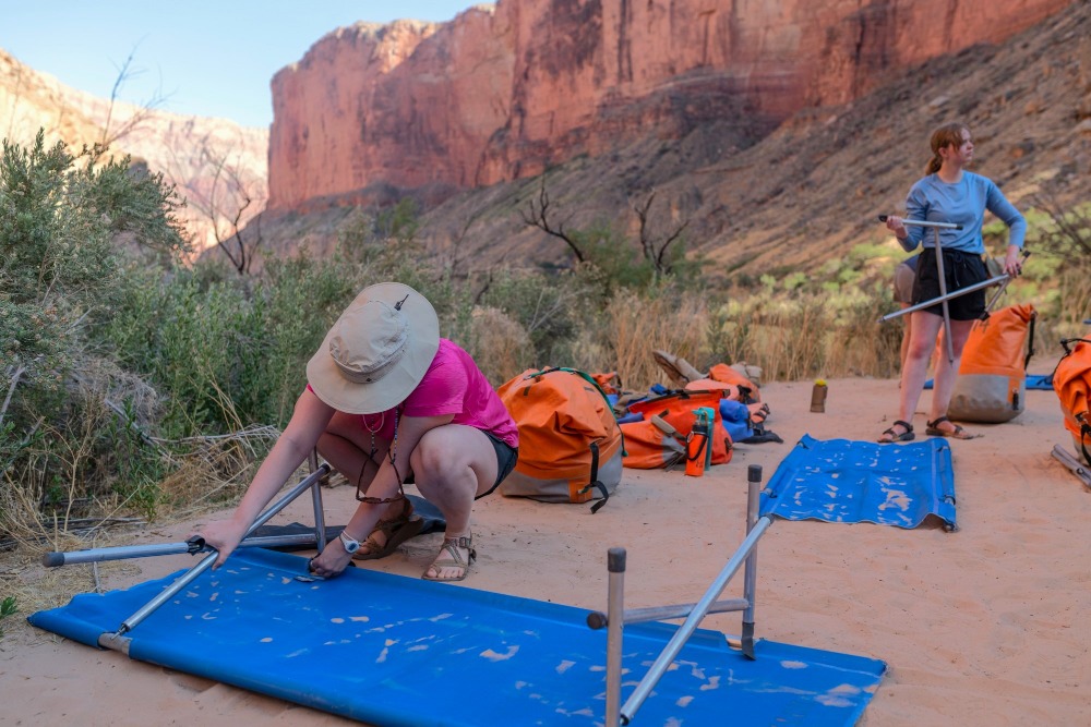 people set up a cot at a sandy campsite along the Colorado River