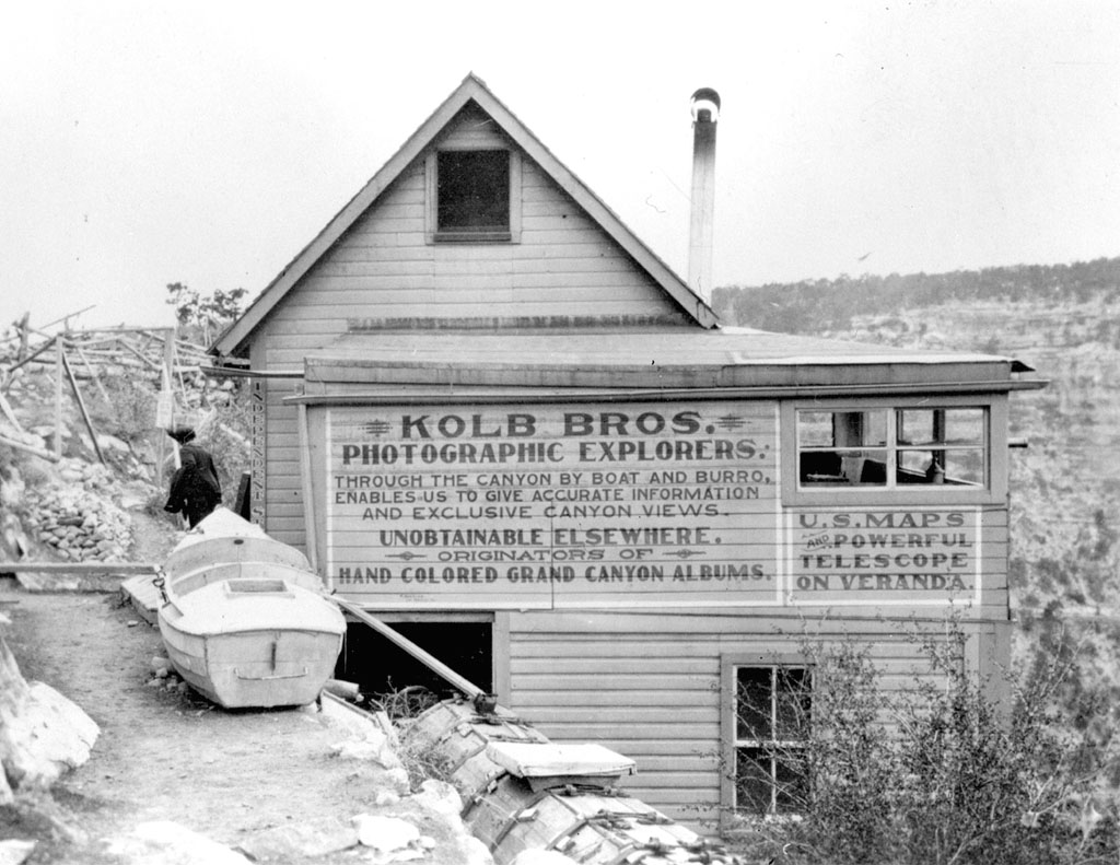 Black and white photo taken in 1912 of the Kolb Brothers photo studio with a river boat in front at Grand Canyon next to the Bright Angel Trail