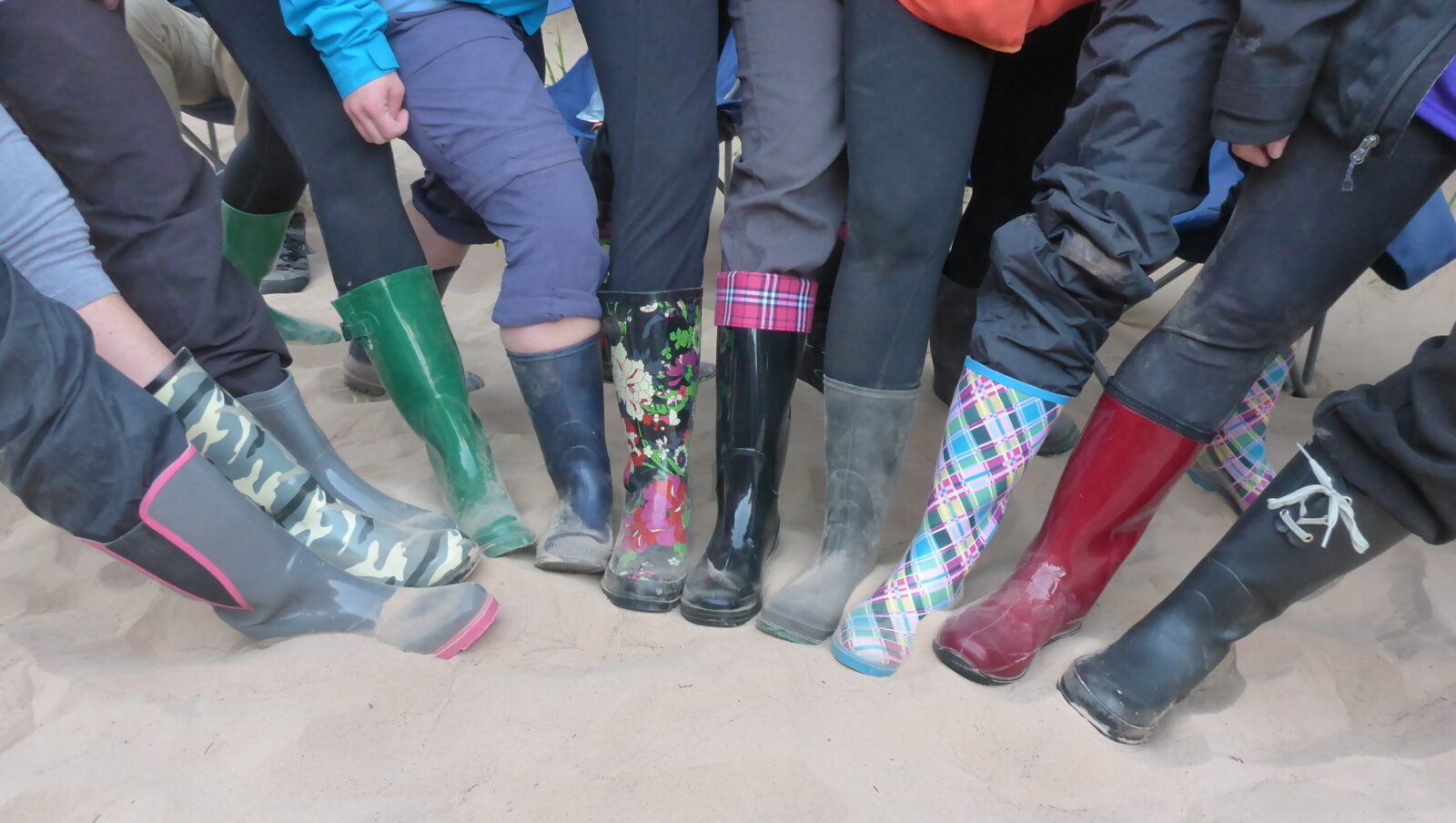 Collection of feet in a line showing off their multi-colored galoshes on a sandy beach. Photo credit: Mary Jo May.