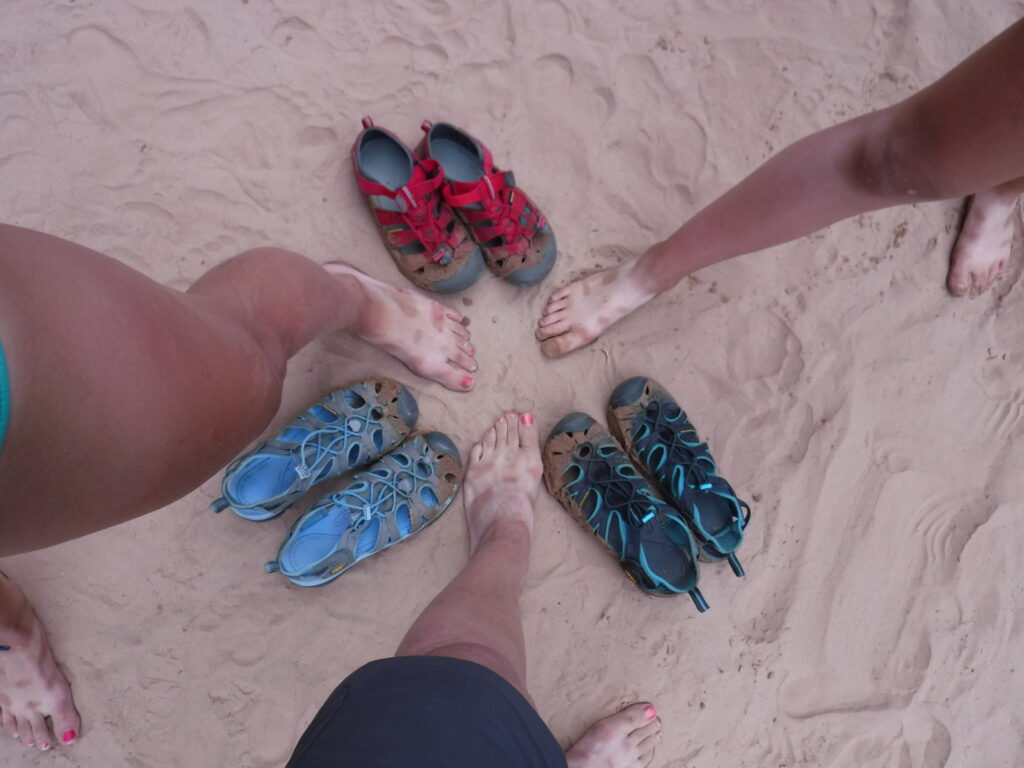 Three people's bare feet in the sand arranged in a circle surrounded by their hiking sandals. Photo credit: Nancy Sanguinetti.