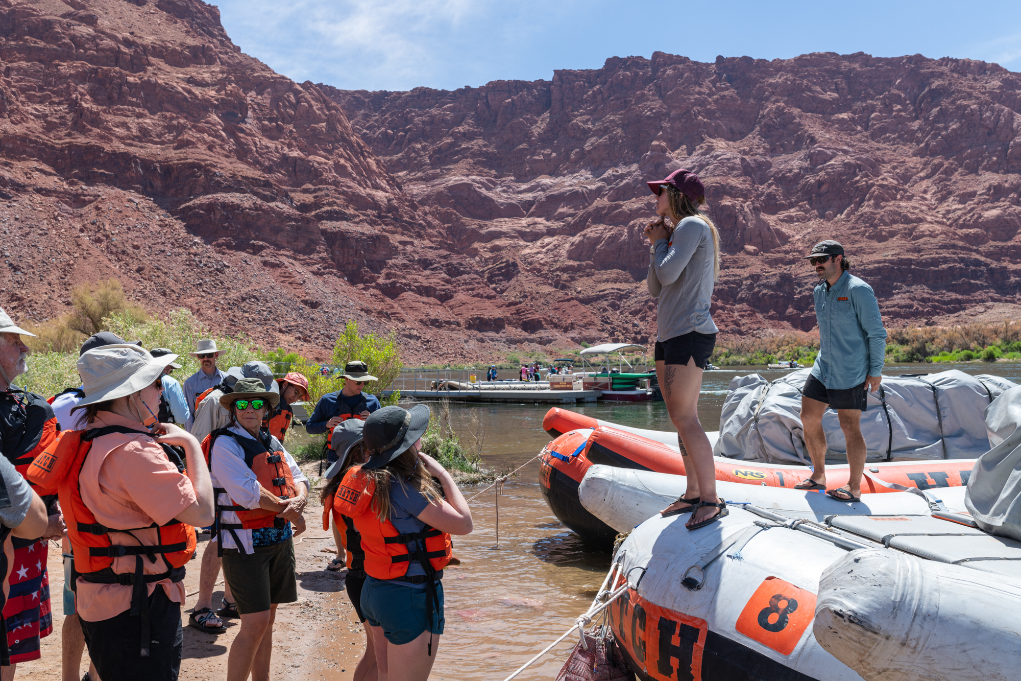 Orientation at Lees Ferry for a river rafting trip through Grand Canyon. People wearing life vests stand in front of the rafts looking up at the guides who are speaking to the group.