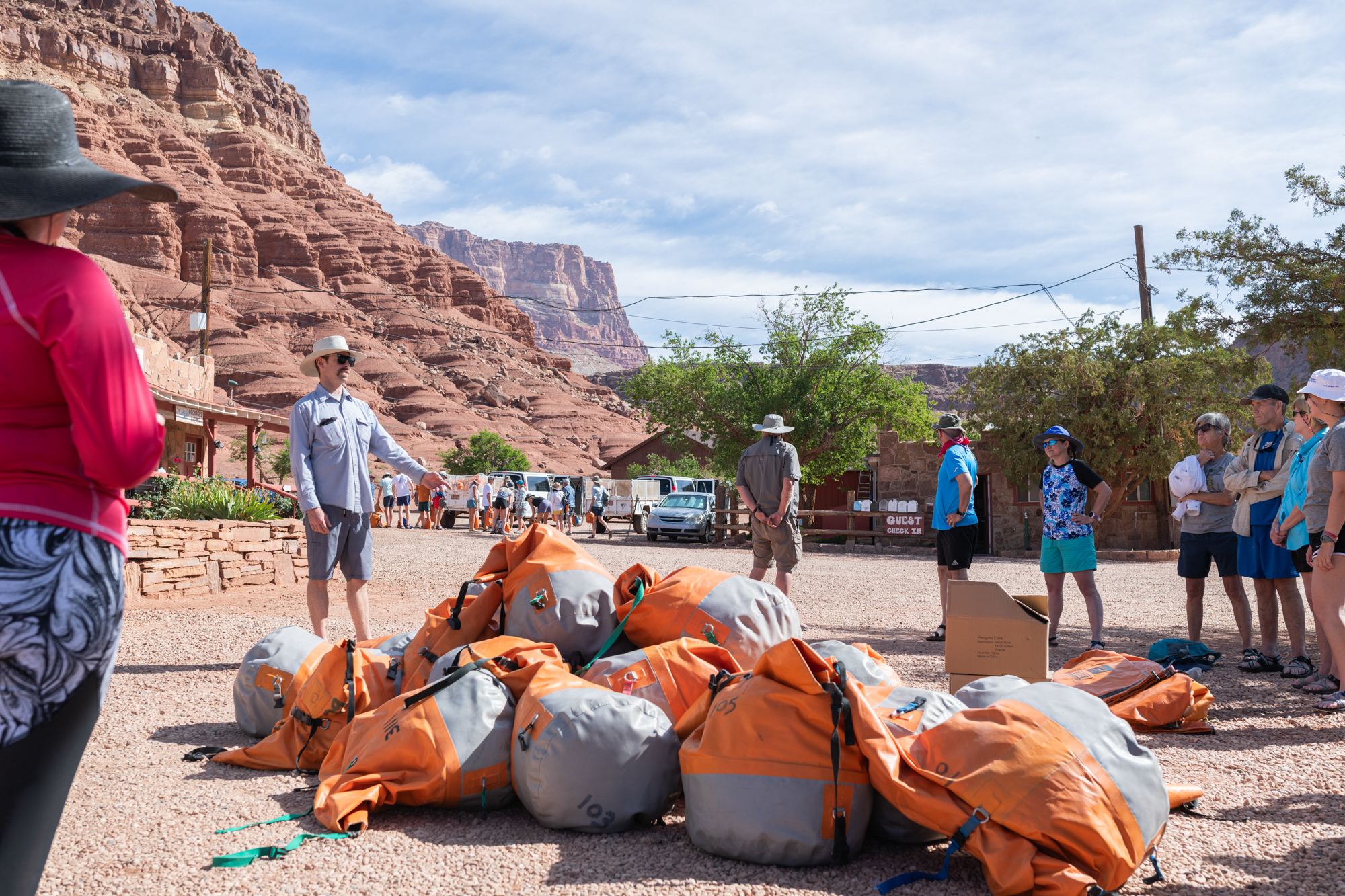 Hatch river rafting orientation outside Cliff Dwellers Lodge. The orientation leader speaks to a group of people while a pile of bright orange, large dry bags is in the center. The lodge and the Vermillion Cliffs are in the background.