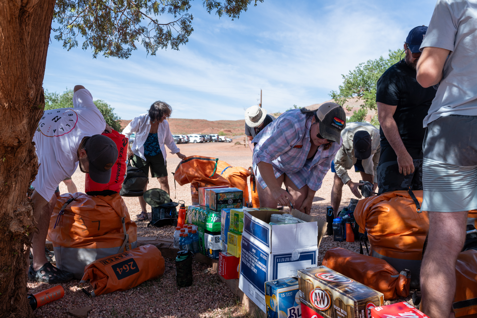 People load their gear into bright orange dry bags and label their drinks under the shade of a tree on a sunny day