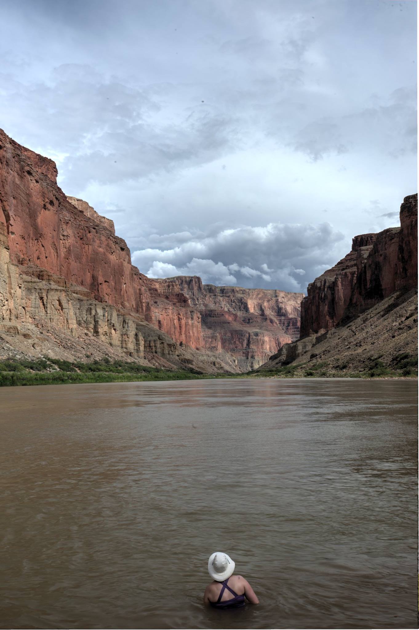 A person stands in the Colorado River looking out at the Grand Canyon where storm clouds characteristic of July and August monsoon season are gathering above the rim. Photo credit: Charles Maudlin.