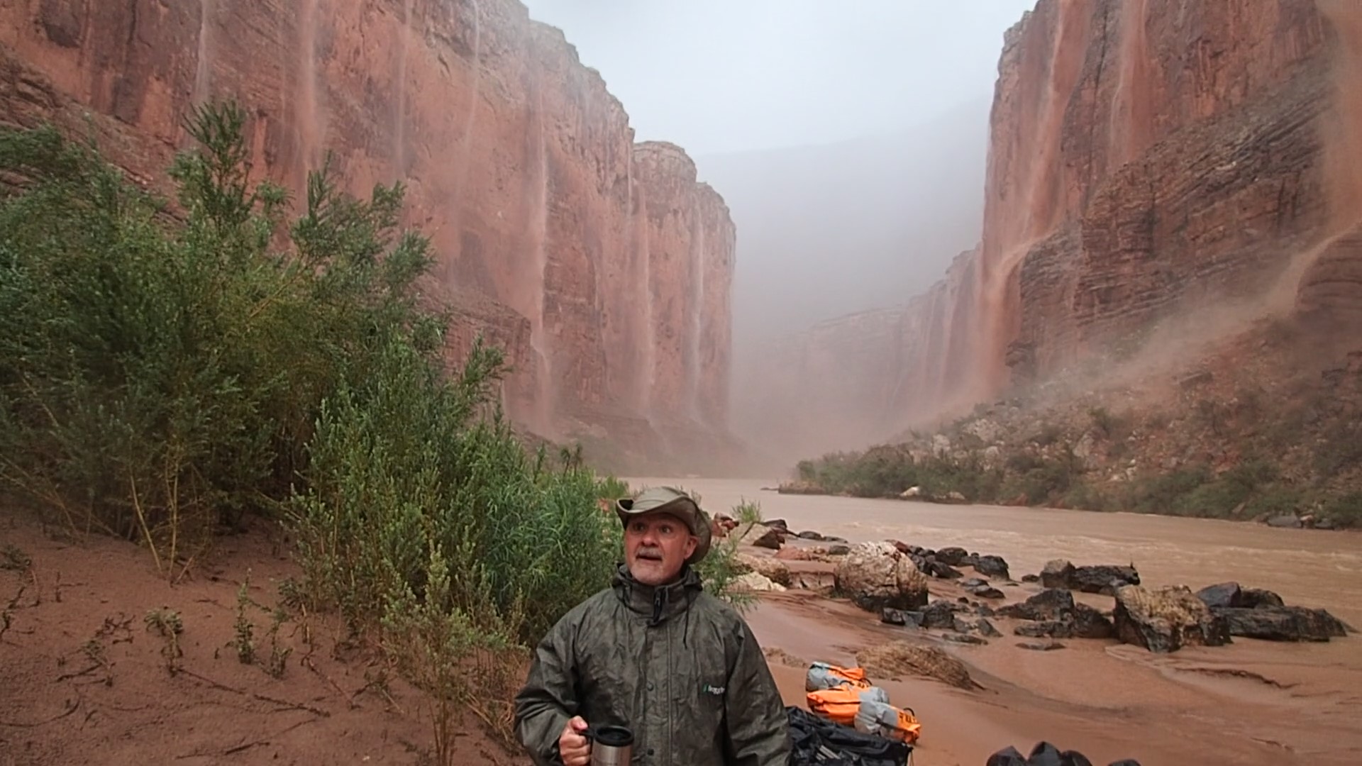 A man stands beside the Colorado River while waterfalls cascade down the cliffsides during monsoons in Grand Canyon. Photo credit: Ken Rein.