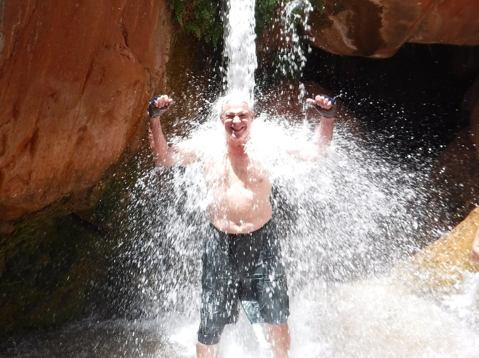 Grand Canyon River Trips are good for health An old man stands under a waterfall, flexing both arms, showing that Grand Canyon river trips are good for your health. Photo credit: Nancy Sanguinetti.